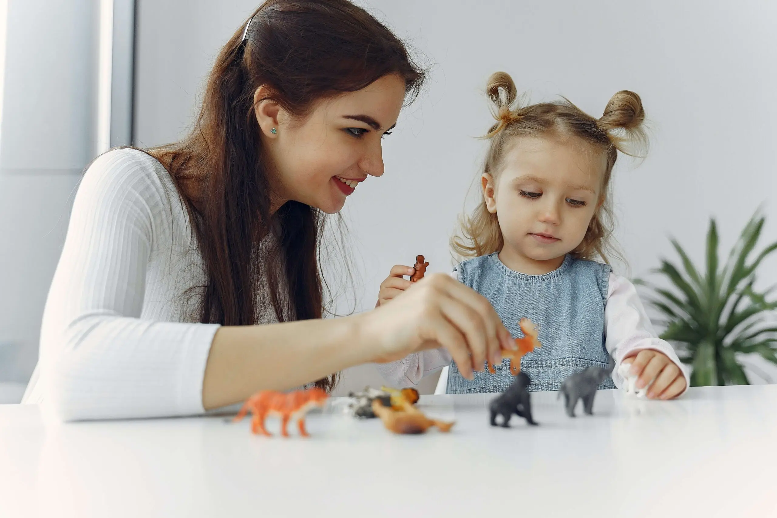 A woman and small female child in a denim jumper play with animal figurines at a table