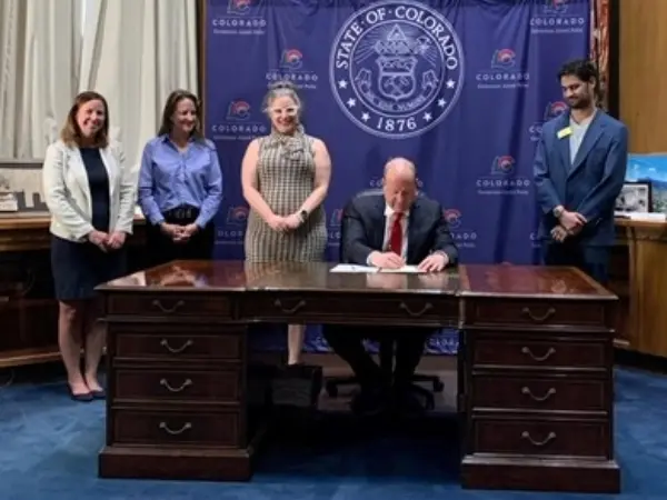 Three women and one man stand behind the governor of colorado as he signs a bill at a desk