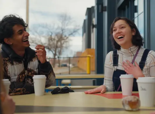 Young people in frame sit outside around a table with other off-screen people while talking and laughing.