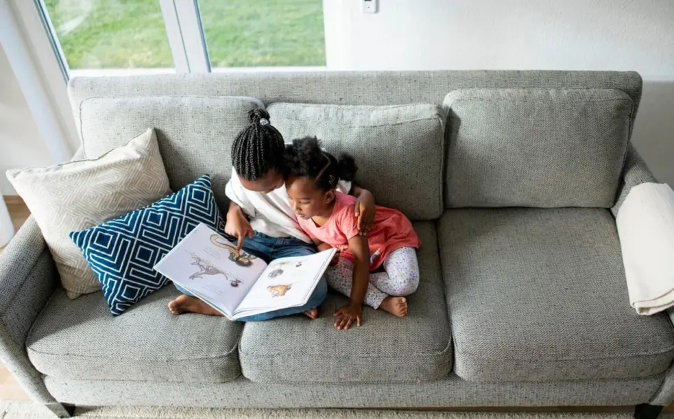 two young sisters sit on couch reading a book together