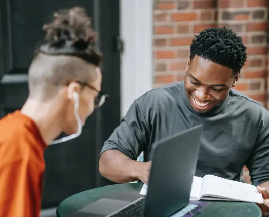 Two college students sit around a table that has a laptop and a notebook.