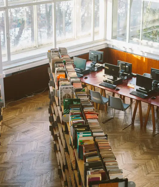 Aerial view of a row of bookshelves next to multiple personal computers inside of a library with many windows.