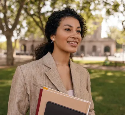 Young woman stands on a lawn wearing a suit and holding folders and a notebook in her hand.