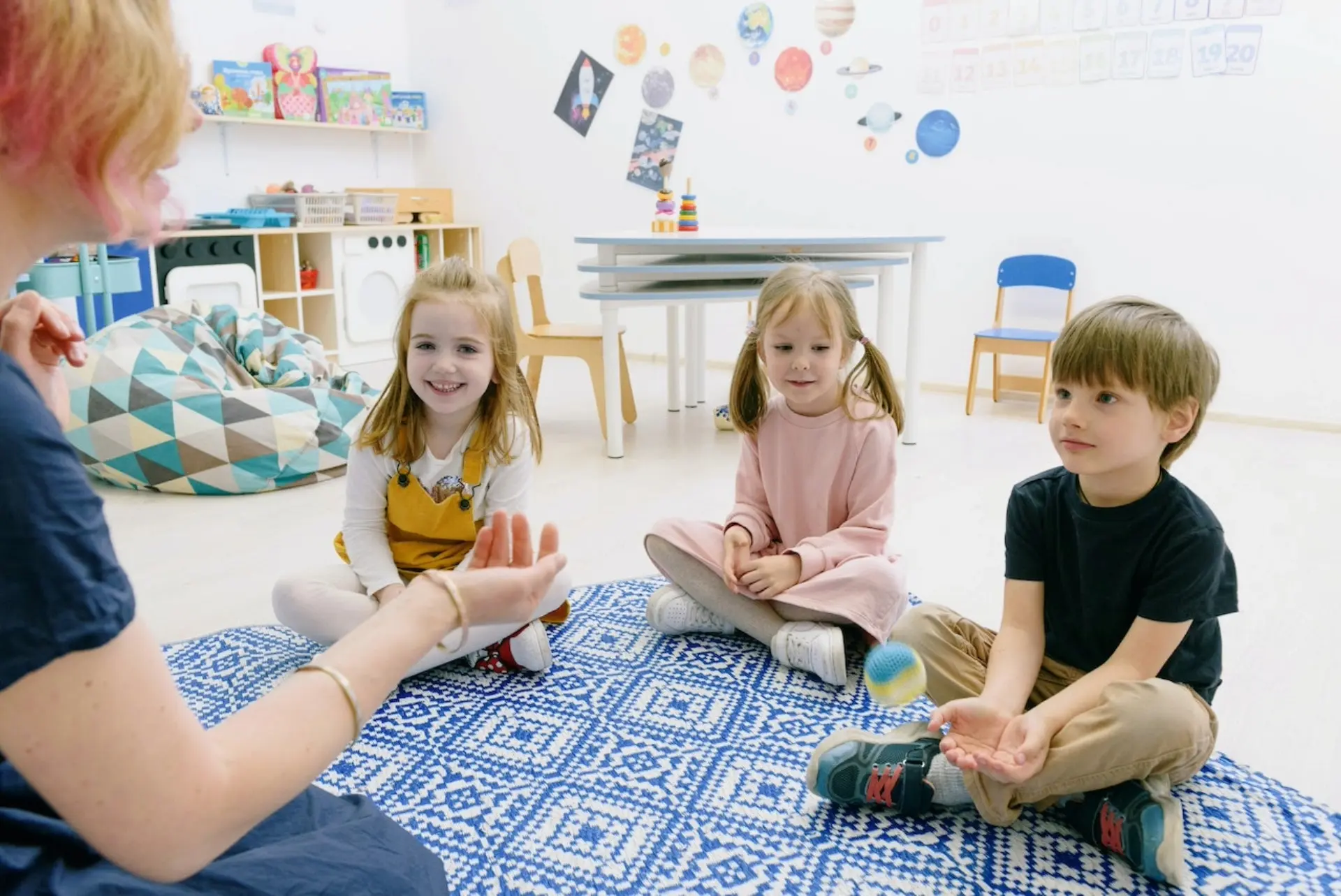 Three preschool children sit on a blue and white carpet while their teacher talks to them