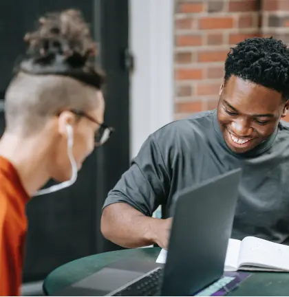 Two young men sit at a table with computer and notebook