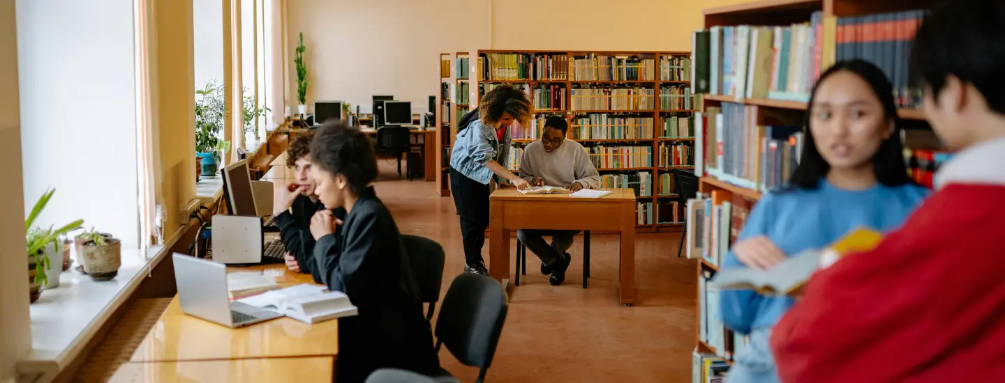 Multiple groups of two college students are shown discussing books in a library.