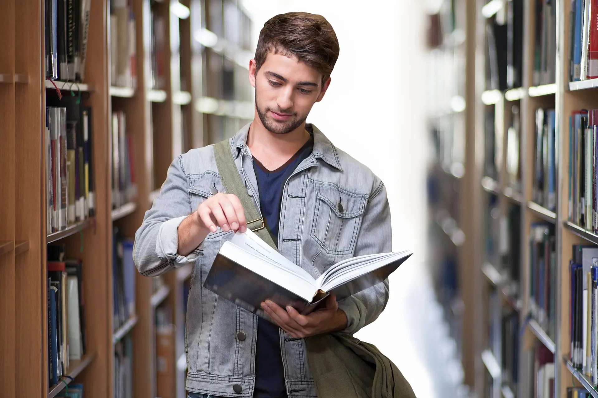 A college age male student in a denim jacket reading a book at the library