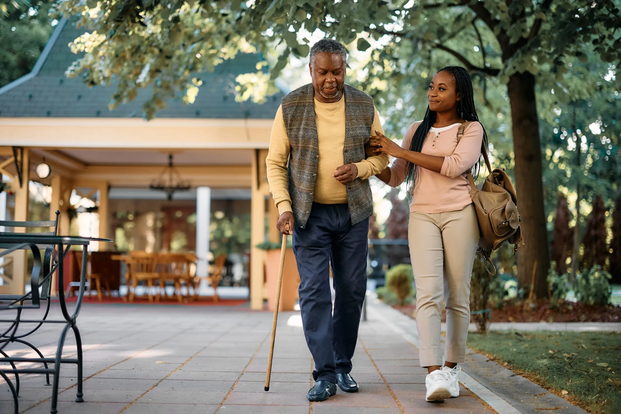 a young woman supports an elderly man walking with a cane through an outdoor sitting area
