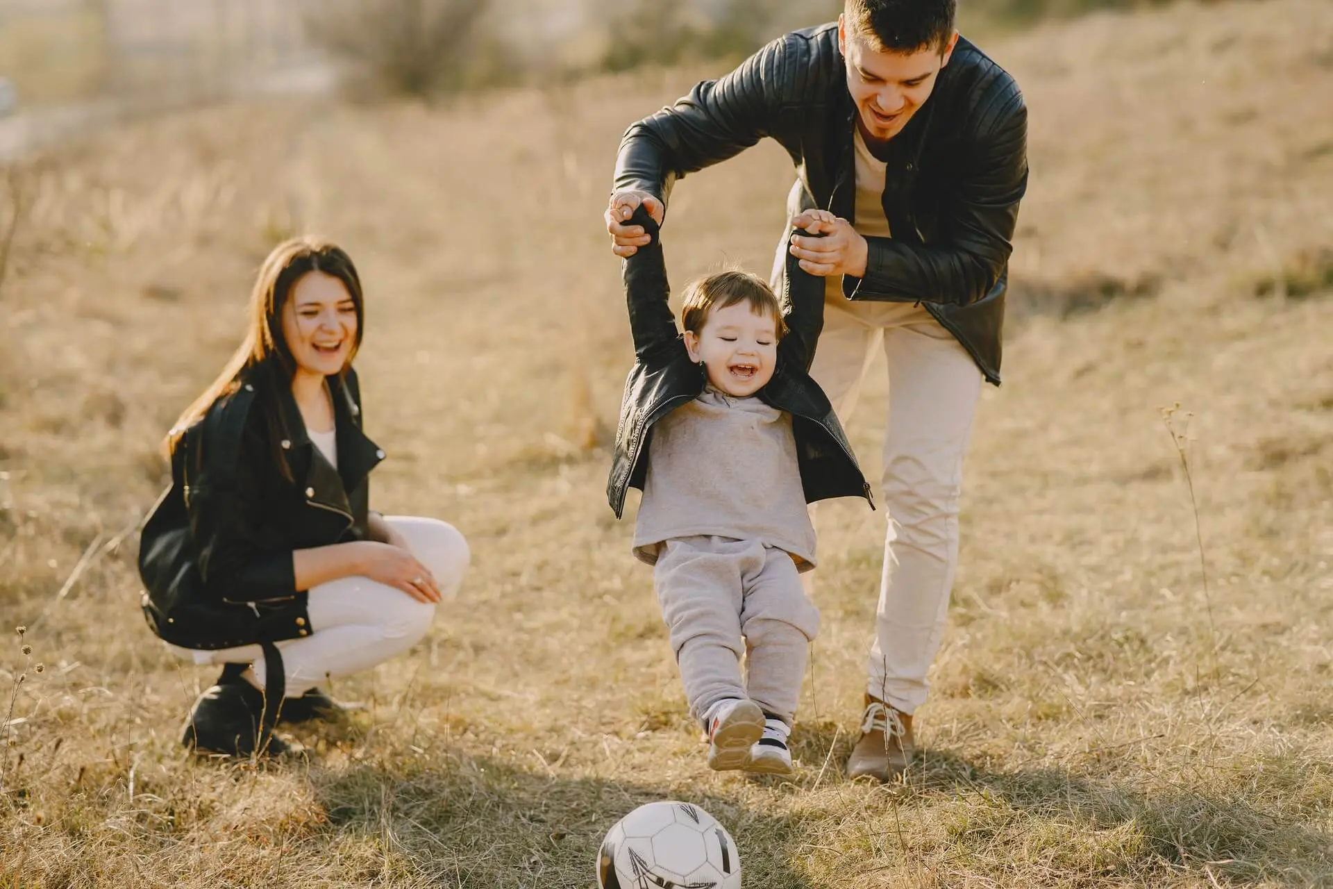 Family of three wearing leather jackets play in a field with a soccer ball while the father holds the toddler by the arms.