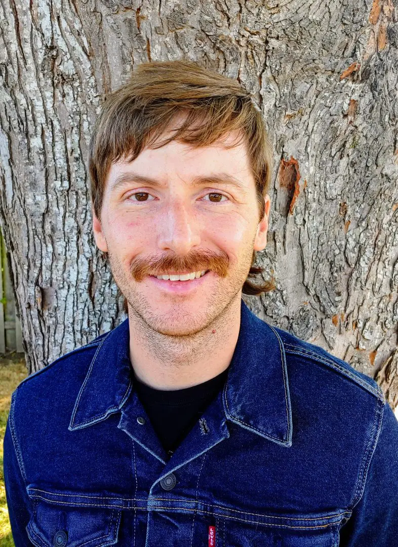 A man in a denim Levi's jacket poses for a headshot in front of a tree