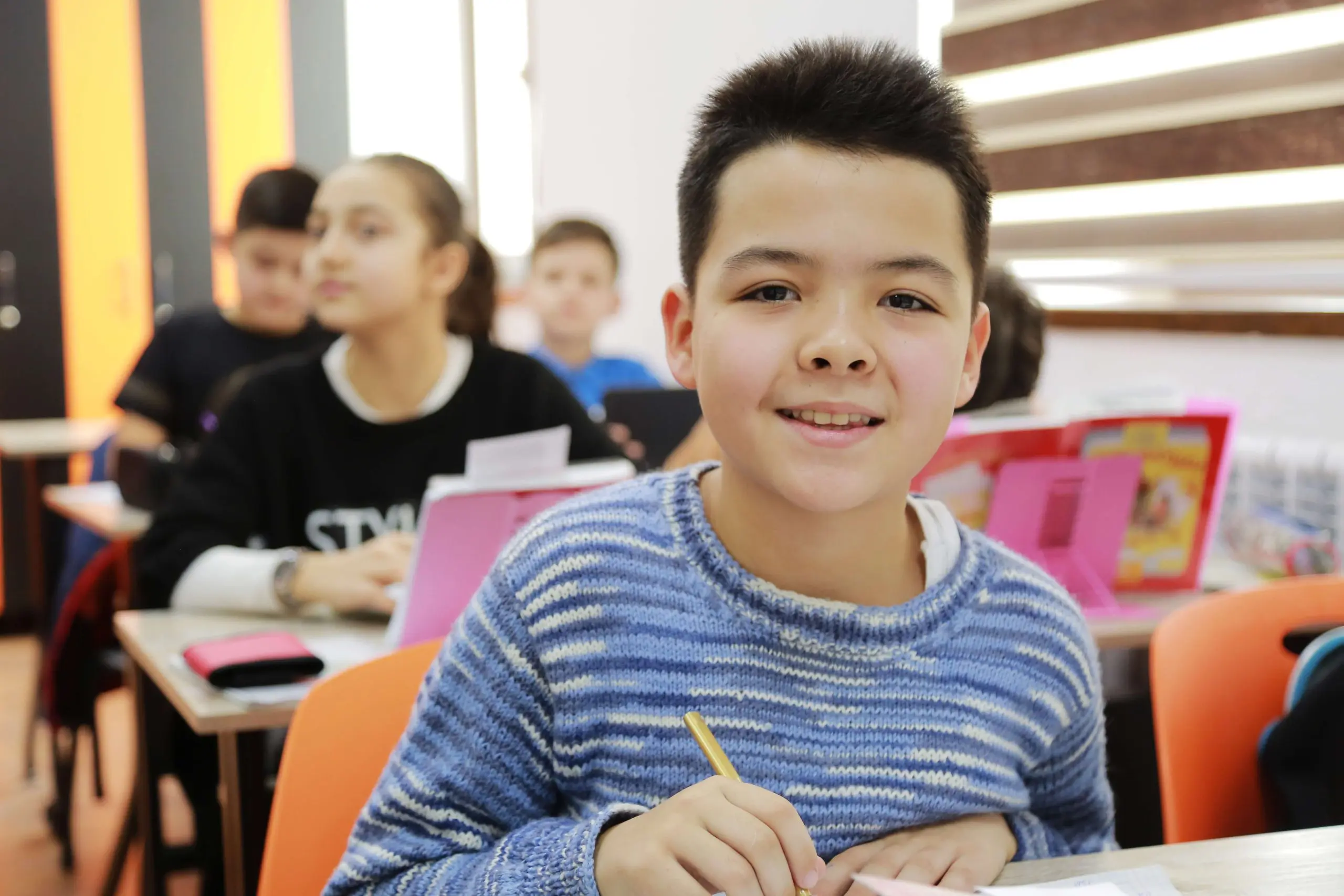 Elementary students in a classroom write and draw while using images in reference books. A boy in center frame smiles at the viewer.