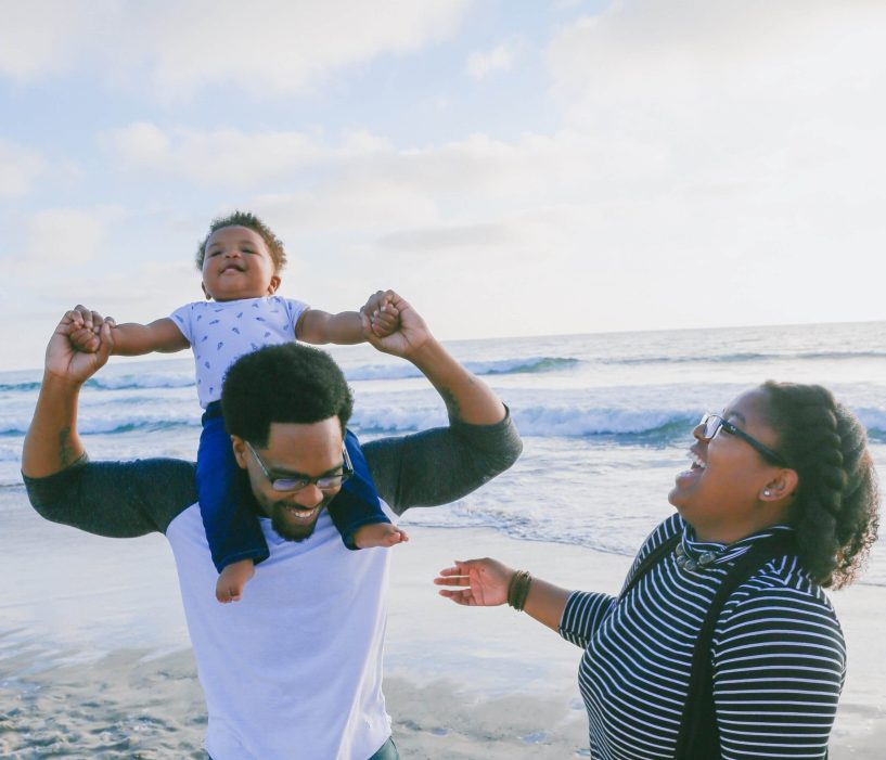 Baby receives piggyback ride on the beach from father while mother watches and laughs.