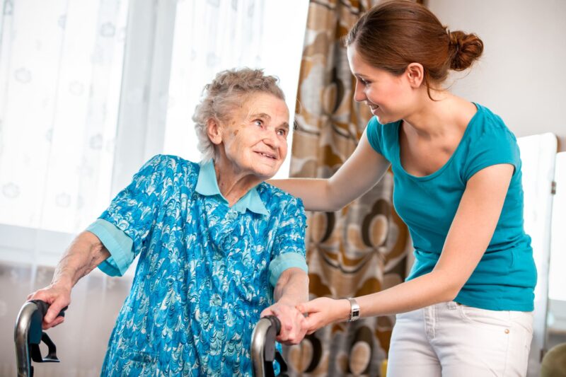 Image of an elderly woman with a walker being assisted by an aide.